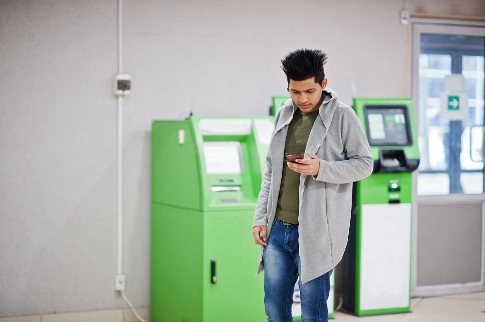 Young stylish asian man with mobile phone against row of green ATM.